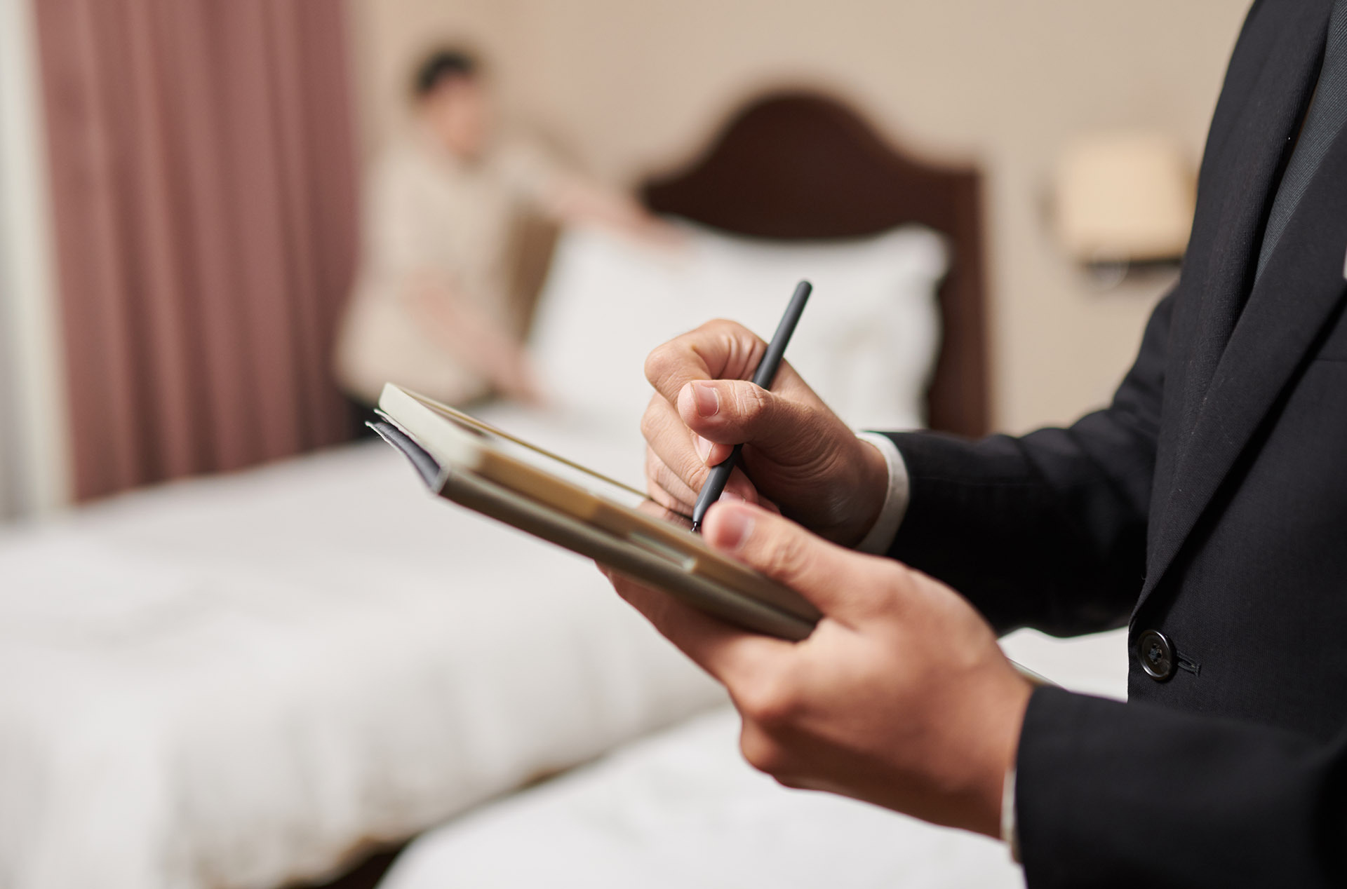 Hotel supervisor taking notes on a notepad while a housekeeper prepares a guestroom bed, highlighting the role of communication in maintaining service standards.
