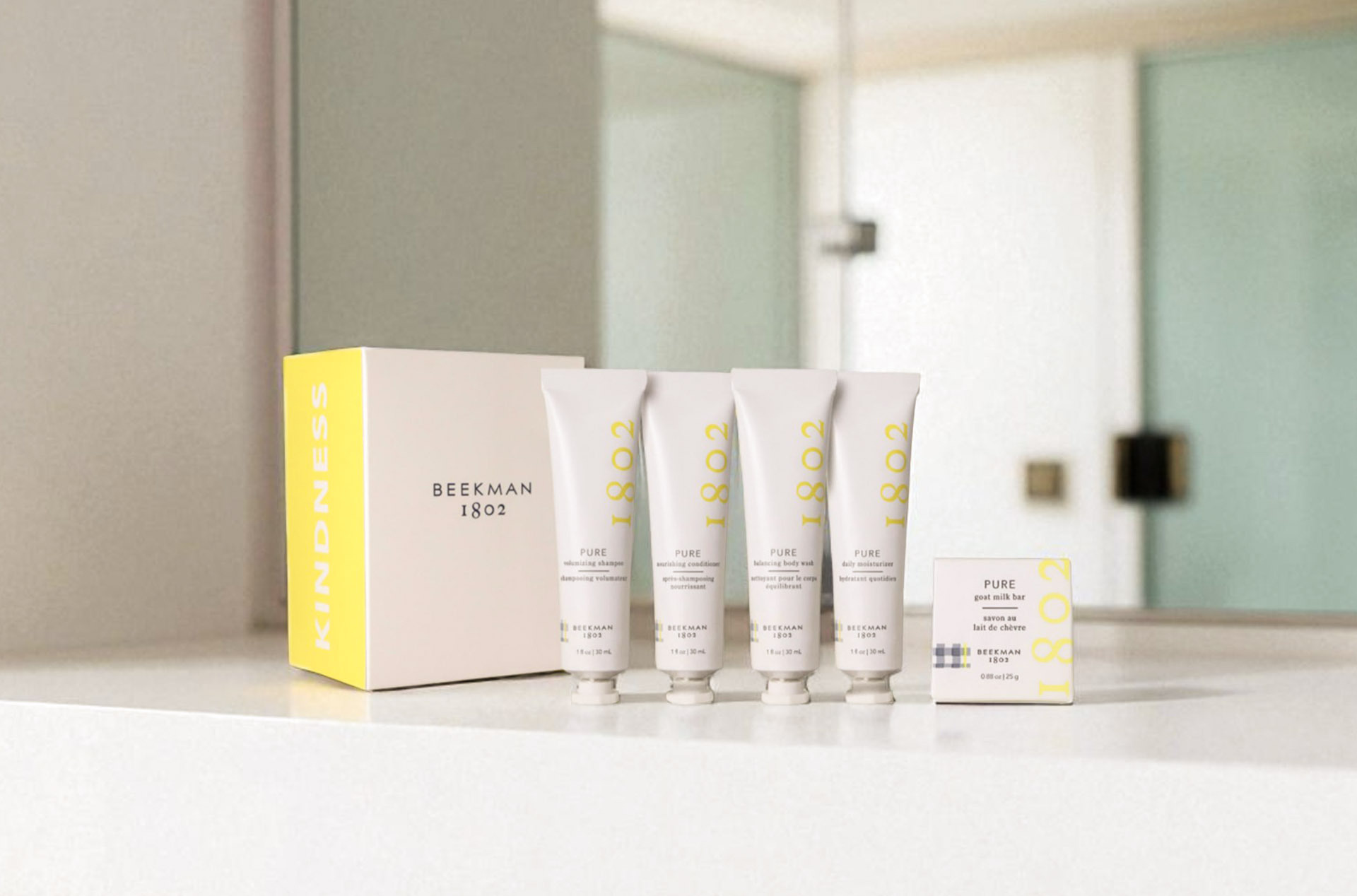 Beekman 1802 Pure amenity kit with travel-size shampoo, conditioner, body wash, moisturizer, and soap displayed on a vanity